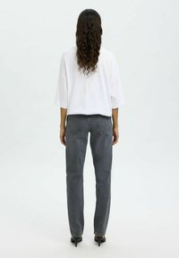 Mujer con el pelo largo y rizado que lleva una camisa blanca holgada, vaqueros negros de pierna recta y zapatos negros de tacón, de pie de espaldas.