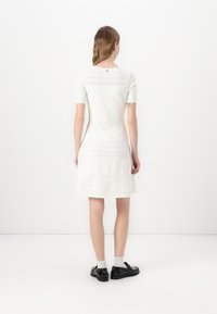 White knitted dress with short sleeves, textured patterns, A-line shape, and a round neckline. Back features a small button detail.
