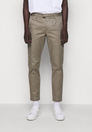 Man draagt beige slim-fit broek, witte T-shirt, witte sokken, witte sneakers en een karabijnhaak vastgeklikt aan een riemlus.