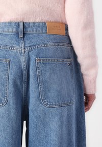 Jeans de mezclilla azul claro con una cinturilla alta y dos bolsillos traseros. Una etiqueta de cuero marrón muestra "TOMMY HILFIGER". Se ve la manga de un suéter rosa suave.