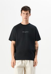 Ung mann med kort mørkt hår, iført svart Quiksilver t-skjorte og lysebeige bukser, som står mot en ensfarget hvit bakgrunn.