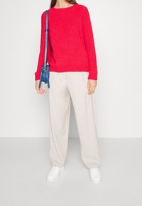 Rode gebreide trui met geribbelde manchetten, gecombineerd met losse beige broek en witte sneakers. Een kleine blauwe schoudertas wordt over de schouder gedragen.