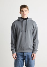 Grijze pullover hoodie met aanpasbare trekkoorden, zachte textuur en subtiele branding op de borst, gecombineerd met lichtblauwe jeans.