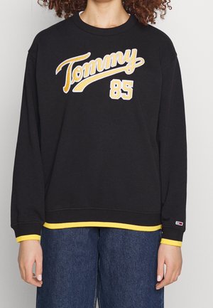 Zwarte sweatshirt met "Tommy" en "85" in gele borduursel, contrasterende gele manchetten en zoom, relaxte pasvorm, zacht materiaal.
