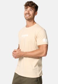Világos bézs színű, rövid ujjú pamut póló, fehér logó grafikával a mellrészen és fehér díszítéssel az ujjon.