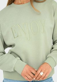 Lichtgroene sweatshirt met het woord "ENJOY" geborduurd op de voorkant, gedragen door een persoon met gekruiste handen, die een zilveren ring en een spijkerbroek draagt.