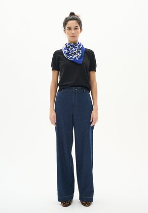 Femeie în picioare purtând un top negru cu mâneci scurte, blugi albaștri cu talie înaltă și pantaloni largi, un fular albastru cu alb cu model și pantofi cu imprimeu de leopard.