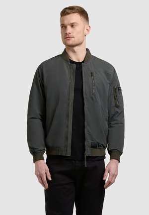 Mladý muž s krátkymi vlasmi, oblečený v tmavozelenej bomber bunde cez čierne tričko, stojaci pred jednoduchým svetlým pozadím.