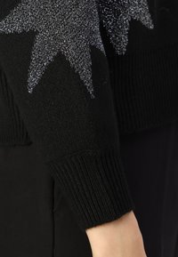 Black knit sweater featuring a ribbed cuff and metallic silver star pattern across the upper sleeve, showcasing texture and contrast in design.