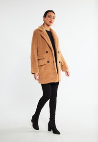 Tan, falsk pels dobbeltbrystet frakke med sorte knapper og sidelommer, båret over en sort top og sorte leggings, parret med hæle støvler.