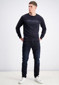 Felpa nera con logo testurizzato, scollo tondo e maniche lunghe, abbinata a jeans blu scuro e sneakers bianche. Design semplice.