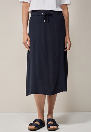 Marineblaue lange Rock mit strukturierter Oberfläche, Kordelzugbund und seitlichen Taschen, kombiniert mit dunklen Sandalen.