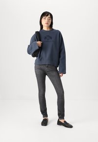 Sweat-shirt bleu marine avec un design graphique sur la poitrine, associé à un jean skinny gris et des mocassins noirs. Le modèle porte un sac noir texturé.
