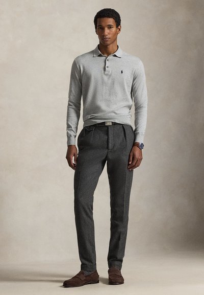 Polo a maniche lunghe di colore grigio chiaro con tre bottoni, abbinata a pantaloni plissettati grigio scuro e mocassini marroni. Lo sfondo è neutro e morbido.