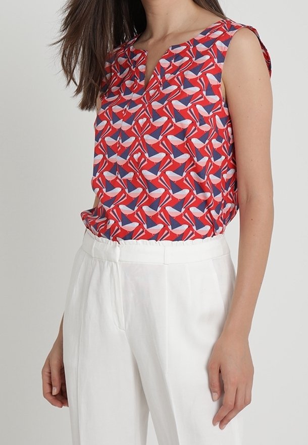 Rote, gemusterte ärmellose Bluse mit V-Ausschnitt, die geometrische Formen in Marineblau und Pink zeigt, kombiniert mit weißen, hochtaillierten Hosen.