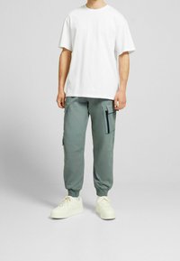 Vit bomullströja som matchas med ljusgröna cargobyxor med sido-fickor och dragkedjedetalj, kompletterat med beige sneakers.