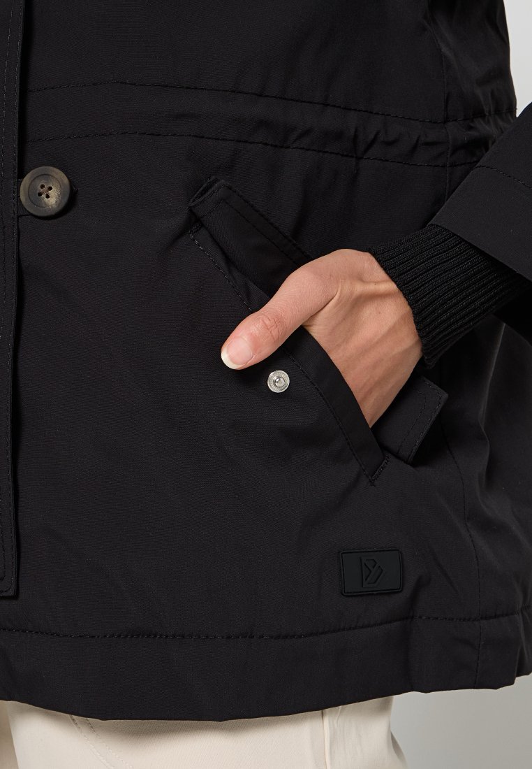 Die schwarze Jacke verfügt über einen strukturierten Stoff, einen Knopfverschluss und eine Seitentasche mit sichtbarem Druckknopf. Logo-Patch auf der unteren linken Seite.