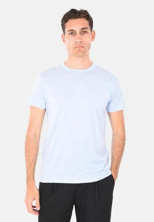 Giovane uomo con capelli corti e scuri, che indossa una maglietta azzurra a maniche corte e pantaloni neri, in piedi su uno sfondo bianco neutro.