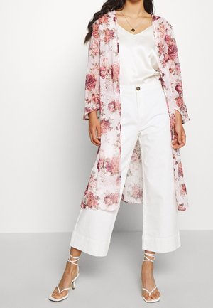 Frau trägt weiße weit geschnittene Hosen, ein weißes Satin-Camisole, einen langen rosa floralen Kimono und weiße Schnür-High Heels, steht vor einem weißen Hintergrund.