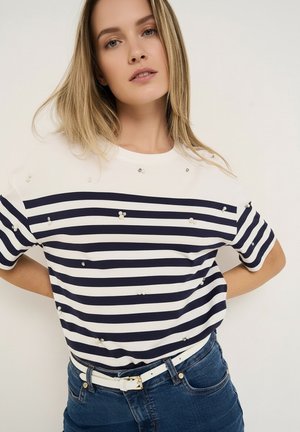 Lyshåret kvinde iført en hvid og marineblå stribet kortærmet skjorte med perlepynt og blå jeans, stående med hænderne på hofterne.
