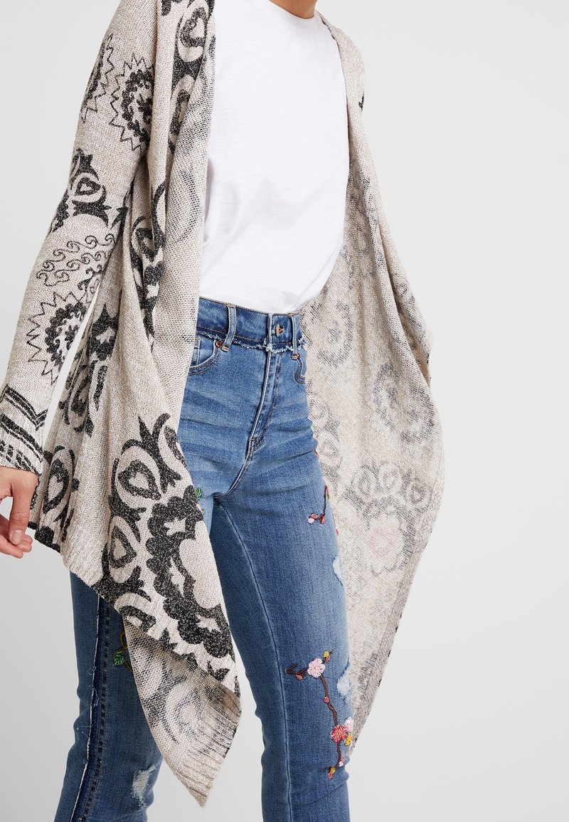 Lichtgewicht, beige cardigan met zwarte gedetailleerde patronen, lange mouwen, open voorkant ontwerp, gecombineerd met blauwe jeans met bloemenborduursels.