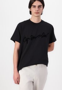 Un T-shirt en coton noir à manches courtes avec un design de logo en relief noir sur le devant. Modèle portant un pantalon de couleur claire.