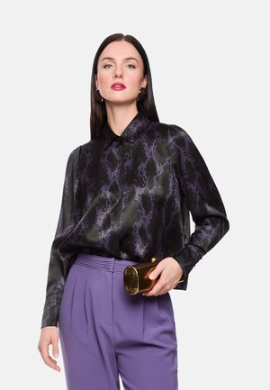 Schwarzes Satinhemd mit lila Schlangenmuster, langen Ärmeln und Kragen; kombiniert mit hoch taillierten lila Hosen und einer goldfarbenen, strukturierten Clutch.