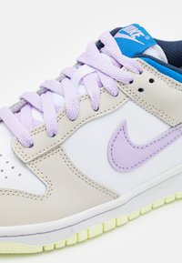 Nike sportcipő fehér bőr felsőrészével, bézs részletekkel, lila fűzőkkel és világoszöld talppal. Figyelemre méltó lila pipa logó az oldalon.