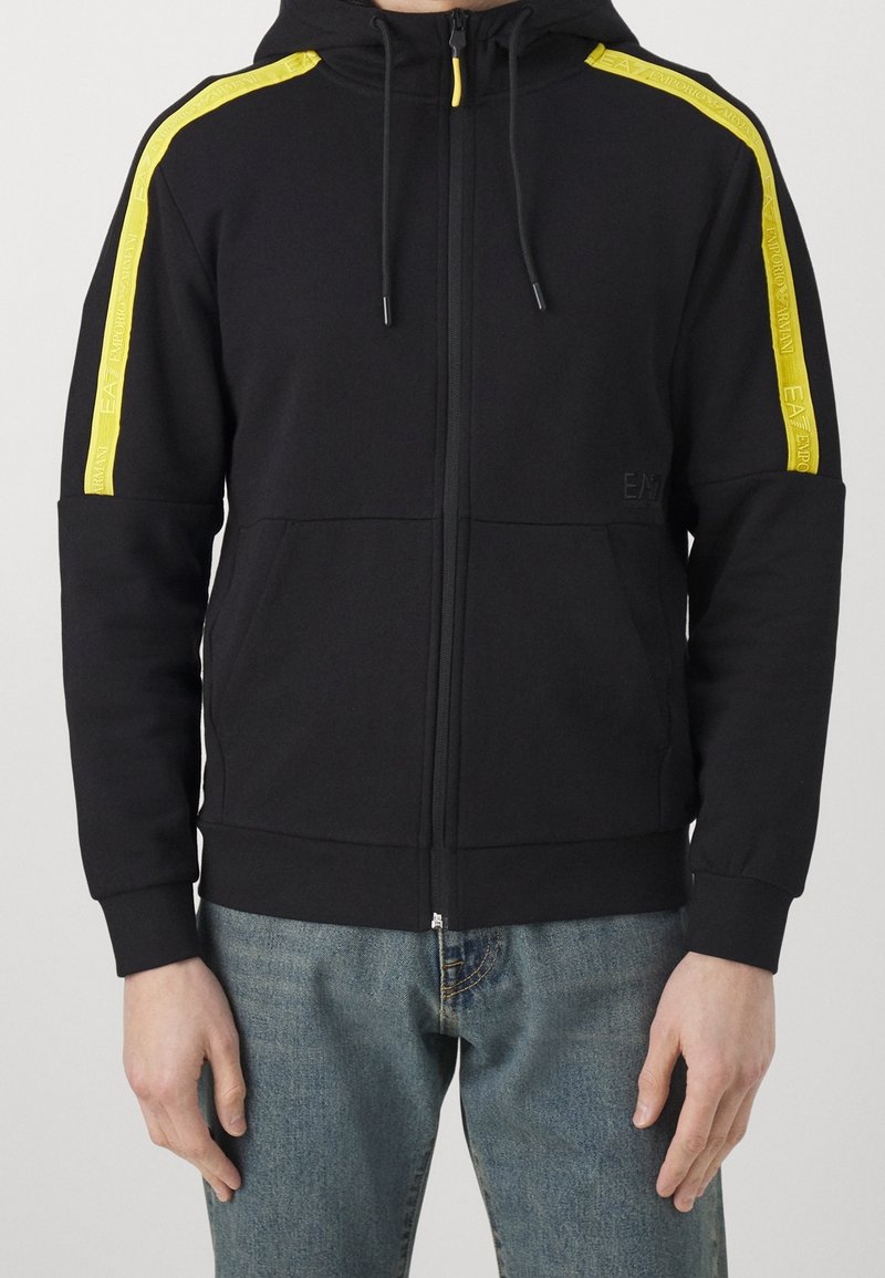 Černá mikina na zip s žlutými pruhy Emporio Armani EA7 na ramenou, klokankové kapsy a modré džíny na stojící osobě.
