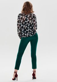 Przezroczysta czarna bluzka z kwiatowym wzorem; zielone dopasowane spodnie; modelka ma na sobie burgundowe szpilki. Widok z tyłu podkreślający szczegóły odzieży.