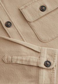 Beige tygskjorta med en texturerad yta, har två fackfickor och två mörka knappar. Detaljer inkluderar en stickad krage och fickkant.