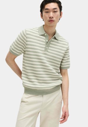 Mann trägt kurzärmliges, grün-weiß gestreiftes Strickpolohemd mit drei Knöpfen und hellbeige Hose vor einfarbigem Hintergrund.