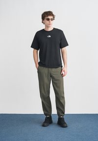 Μαύρο βαμβακερό μπλουζάκι με λευκό λογότυπο, ελαφρώς στενόμενο παντελόνι χακί, και μαύρα παπούτσια. Αξεσουάρ περιλαμβάνουν ανοιχτόχρωμα γυαλιά ηλίου. Απλό φόντο.