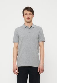 Uomo in piedi di fronte a noi, indossa una polo a maniche corte grigio chiaro e pantaloni neri su uno sfondo bianco semplice.