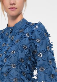 Blått stoff bluse prydet med tredimensjonale blomsterapplikasjoner og paljetter. Har krage og knappeåpning. Teksturt overflate.