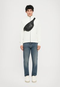 Homem a usar um suéter branquíssimo com fecho, jeans azuis, ténis brancos e uma mochila preta a tiracolo, de pé contra um fundo simples.