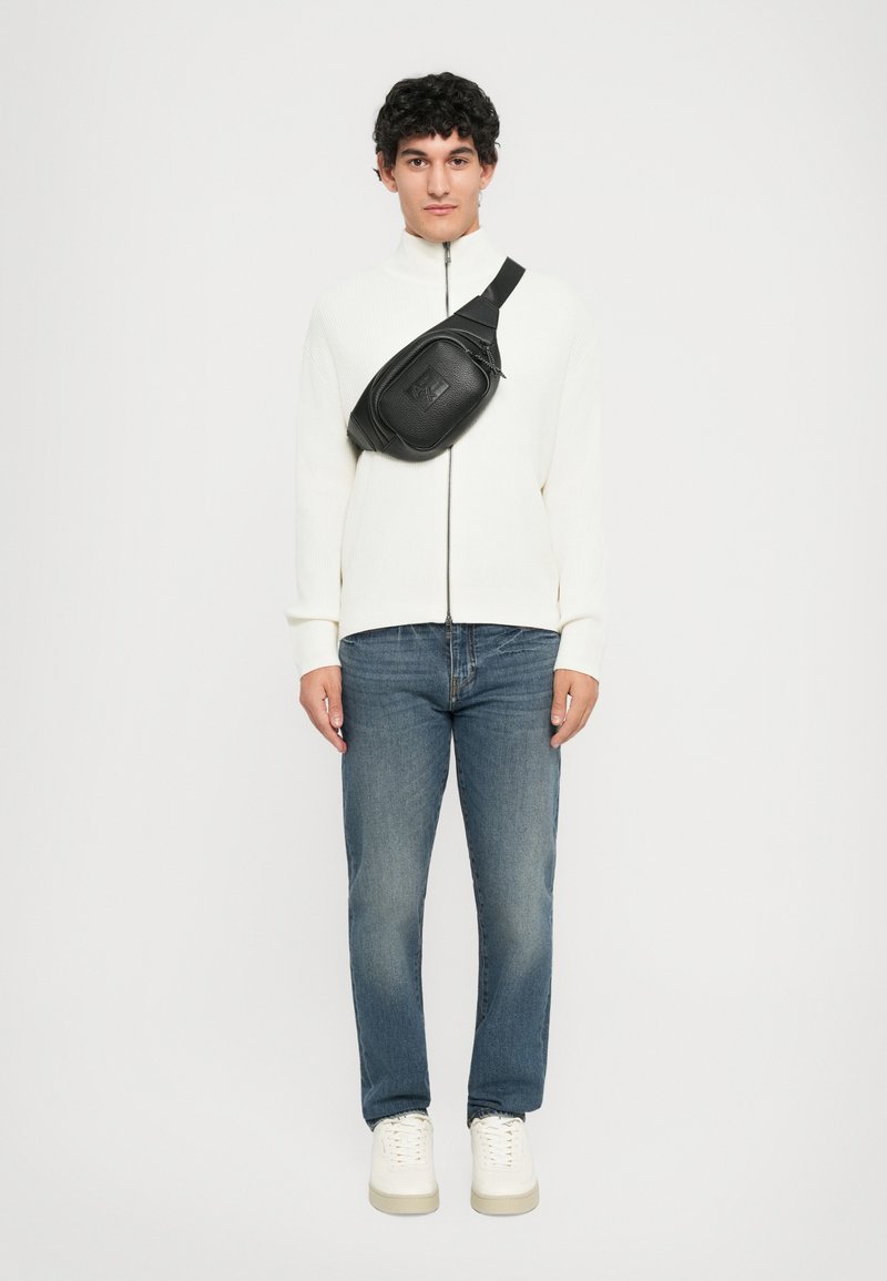 Homem a usar um suéter branquíssimo com fecho, jeans azuis, ténis brancos e uma mochila preta a tiracolo, de pé contra um fundo simples.