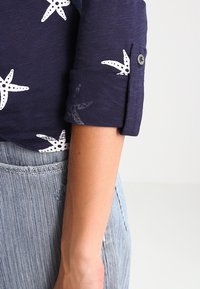 Meripaitoja, jossa on valkoisia meritähtiä, käärityt hihat nappidetaililla, yhdistetty vaaleansinisiin raidallisiin farkkuihin, esittelee kankaan tekstuuria.