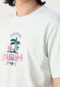 Osoba nosząca białą koszulkę Adidas z zielonym wzorem palmy i różowym napisem "Originals" na piersi, widoczne krótkie ciemne włosy.