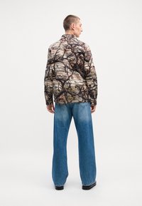 Long-sleeve shirt with a multi-colored, camo-like print, paired with wide-leg, light blue denim jeans and black shoes.