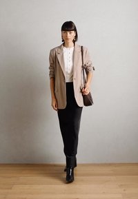 Blazer beige sur une blouse blanche à boutons, associé à un pantalon noir et des bottines. Bijoux simples en accent. Fond neutre.