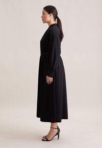 Schwarzes langärmliges Kleid, tailliert mit einem Gürtel, aus glattem Stoff, knöchellang, kombiniert mit schwarzen High Heels.