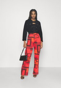 Jaqueta preta curta, blusa preta ajustada e calças largas de cintura alta com estampado geométrico em vermelho e laranja vibrantes. Bolsa preta e sandálias.