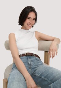 Witte mouwloze top met een hoge halslijn, gecombineerd met lichtblauwe high-waisted jeans en een bruine riem. Model zit op een lichtgekleurde stoel.