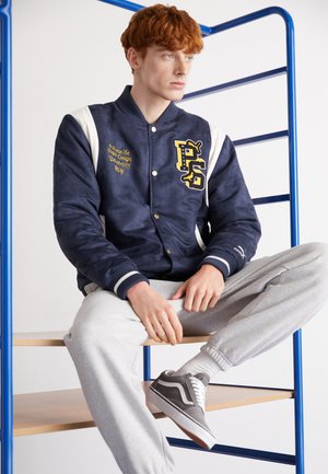 Marine varsityjakke med hvide detaljer, broderet gul logo og trykknapper. Grå sweatpants og grå sneakers fuldender outfittet.