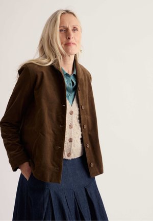Femme aux longs cheveux blonds portant une veste marron, un cardigan crème boutonné, une chemise turquoise et une jupe plissée bleu foncé sur un fond uni.