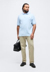 Άνδρας με πλεγμένα μαλλιά, φοράει ανοιχτό μπλε πόλο πουκάμισο και μπεζ παντελόνι, κρατώντας μια μαύρη τσάντα με υφή, στέκεται πάνω σε λευκό φόντο.