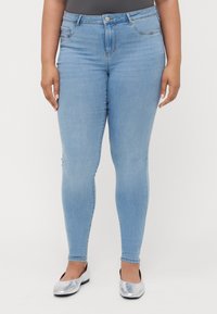 Jeans ajustados de color azul claro con diseño clásico de cinco bolsillos, presillas para cinturón y cierre de cremallera. Llevados con bailarinas plateadas.