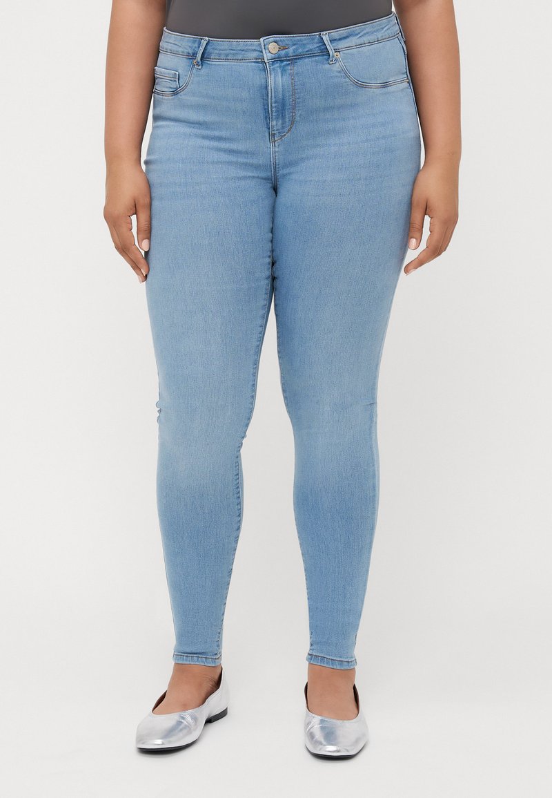 Jeans ajustados de color azul claro con diseño clásico de cinco bolsillos, presillas para cinturón y cierre de cremallera. Llevados con bailarinas plateadas.