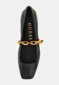 Chaussure plate noire à bout carrée avec une bride en chaîne dorée et le logo "GUESS" à l'intérieur de la semelle, sur un fond blanc.
